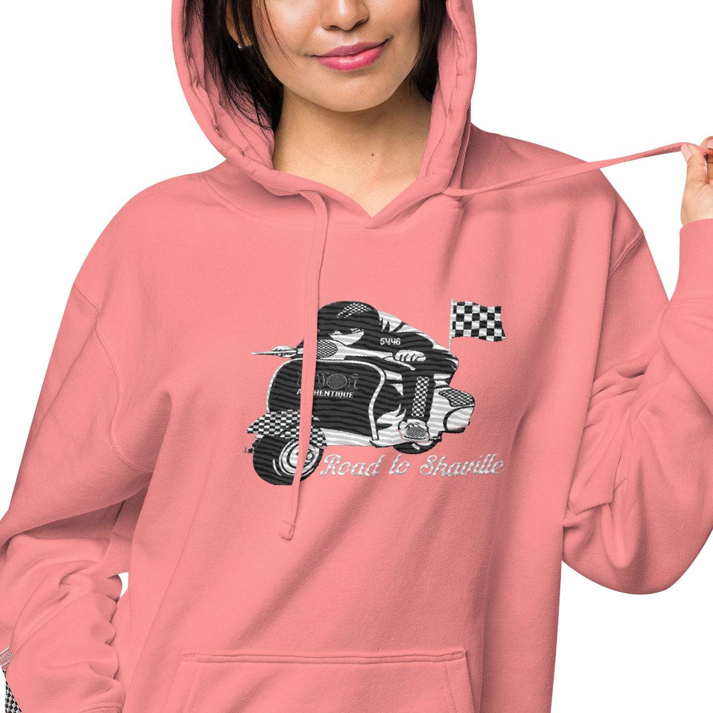 A woman wearing a pink pigment-dyed hoodie with front and sleeve embroidery featuring a vintage moped design and the text 'Road to Skaville'.