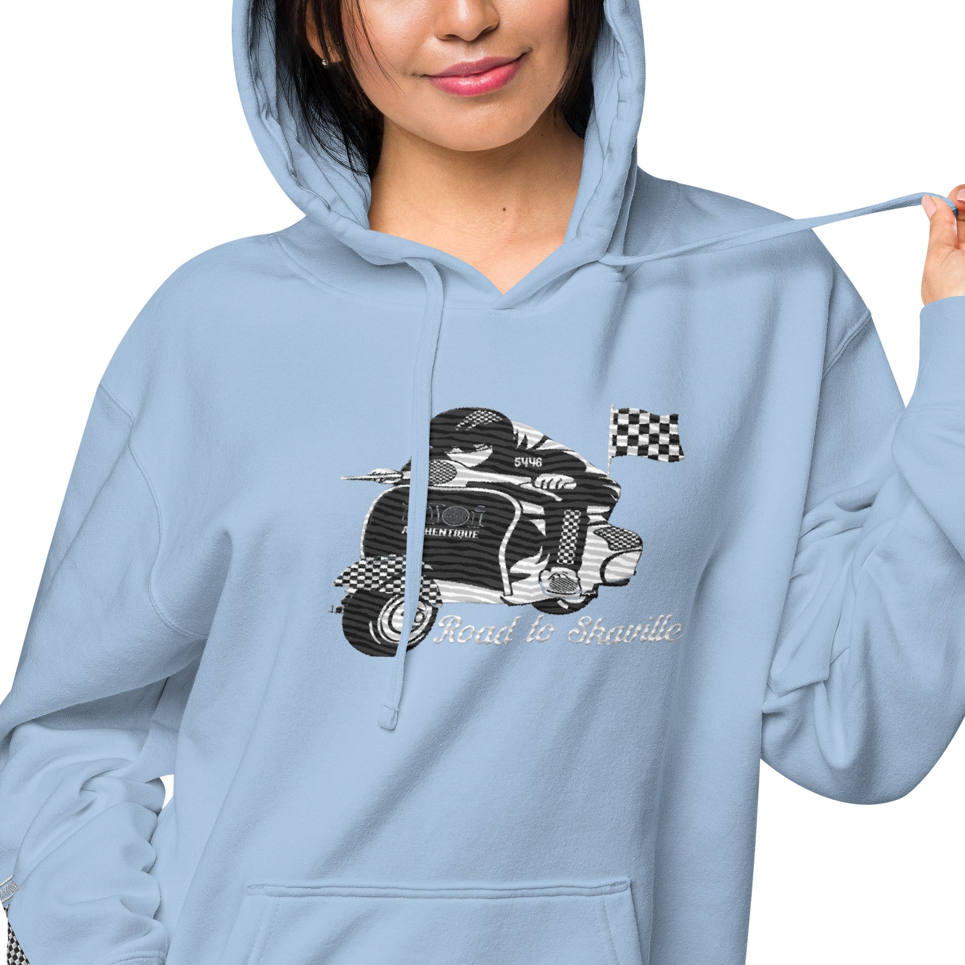 A woman wearing a light blue pigment-dyed hoodie with front and sleeve embroidery featuring a vintage moped design and the text 'Road to Skaville'.