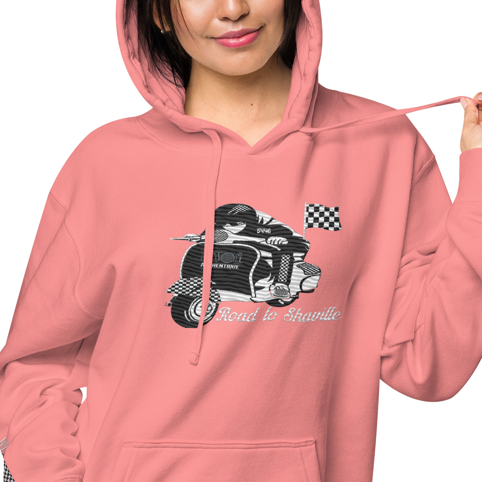 A woman wearing a pink pigment-dyed hoodie with front and sleeve embroidery featuring a vintage moped design and the text 'Road to Skaville'.