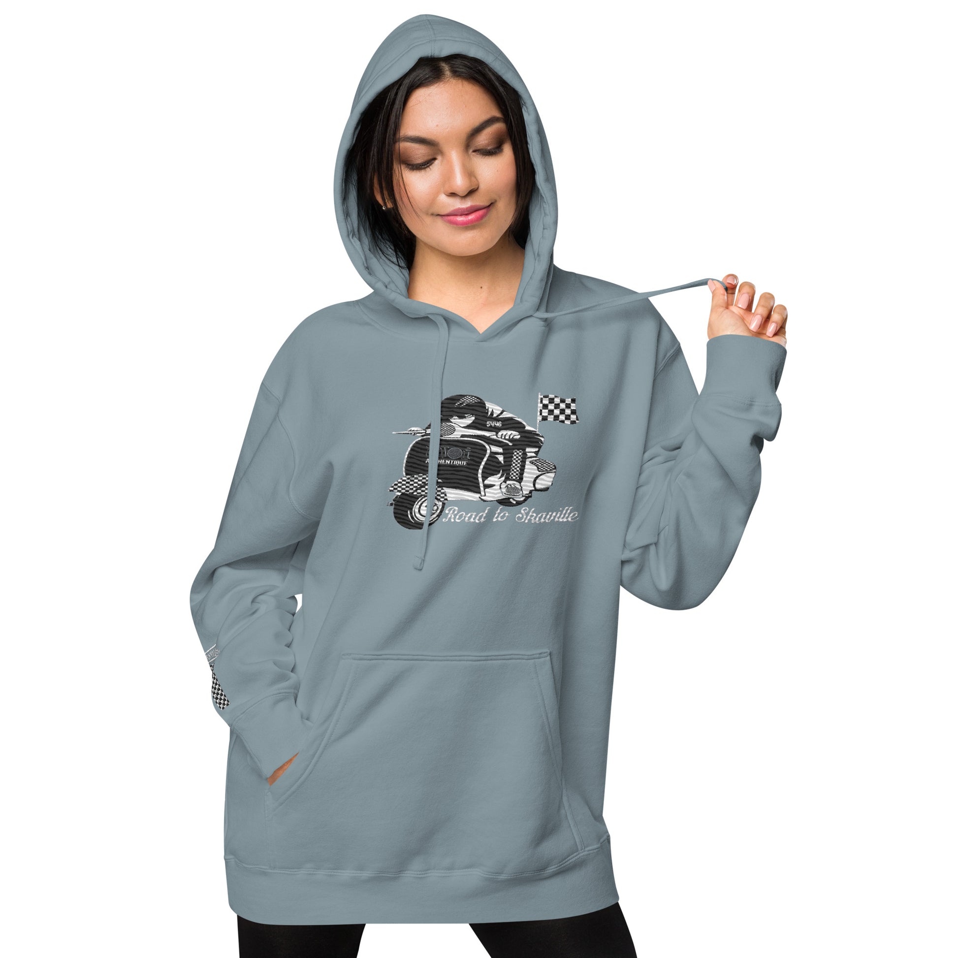 A woman wearing a slate blue pigment-dyed hoodie with front and sleeve embroidery featuring a vintage moped design and the text 'Road to Skaville'.