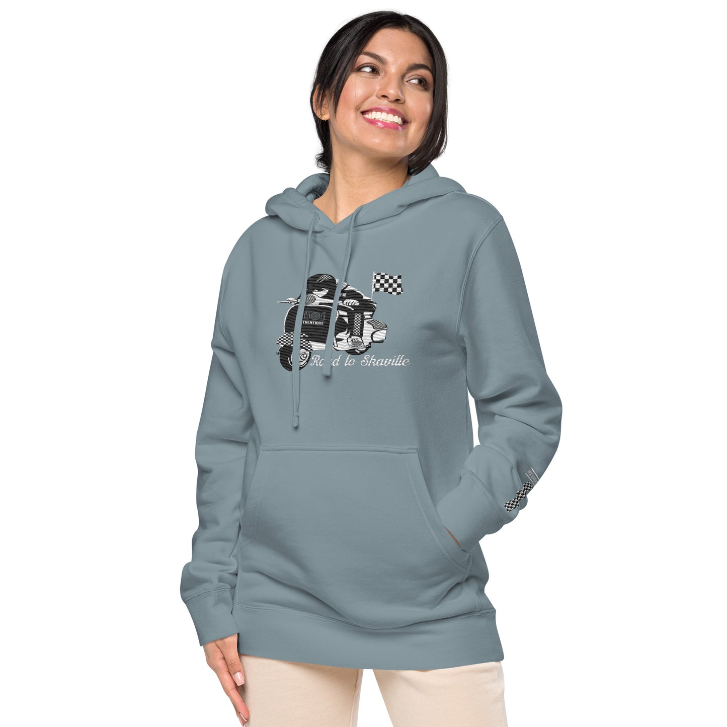 A woman wearing a slate blue pigment-dyed hoodie with front and sleeve embroidery featuring a vintage moped design and the text 'Road to Skaville'.