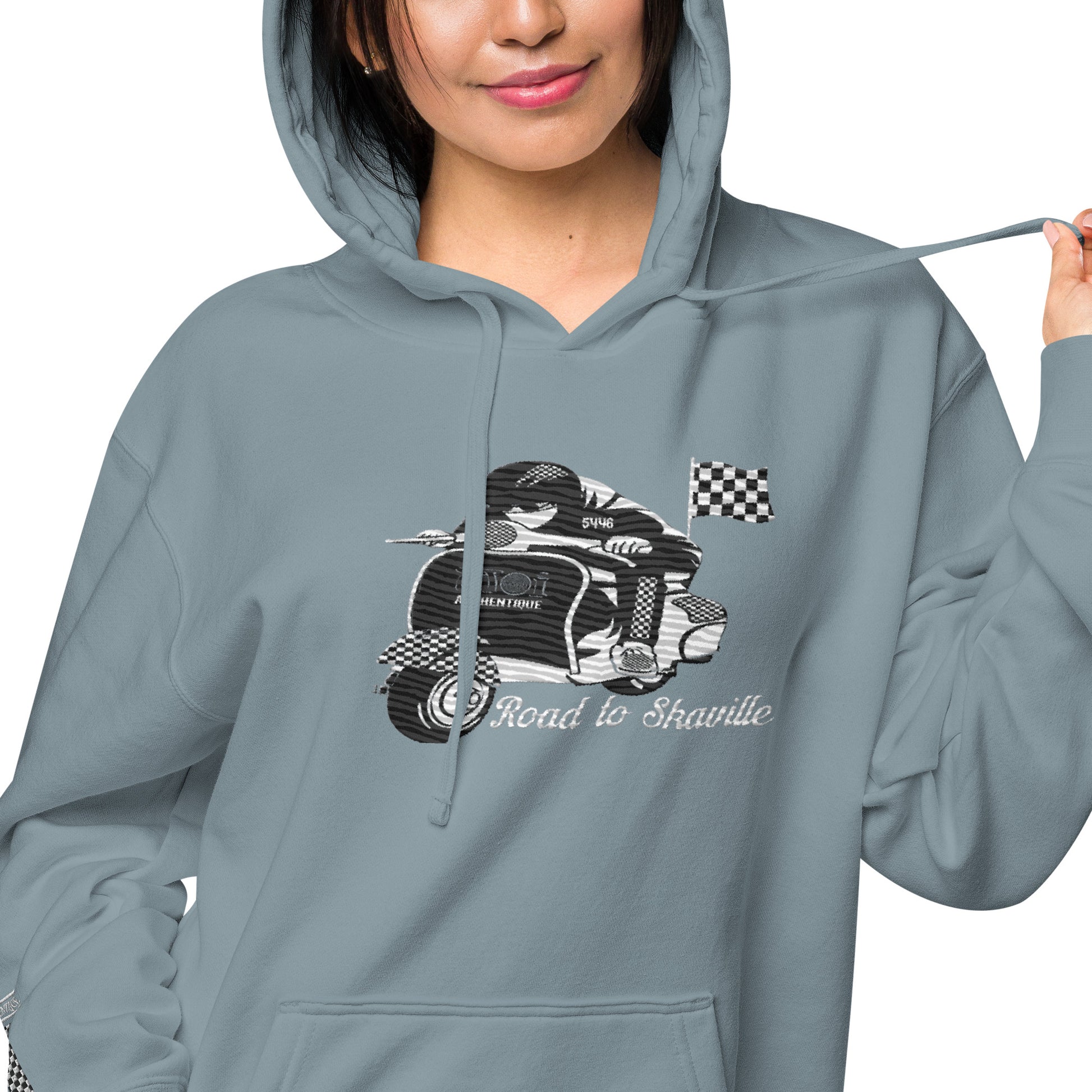 A woman wearing a slate blue pigment-dyed hoodie with front and sleeve embroidery featuring a vintage moped design and the text 'Road to Skaville'.