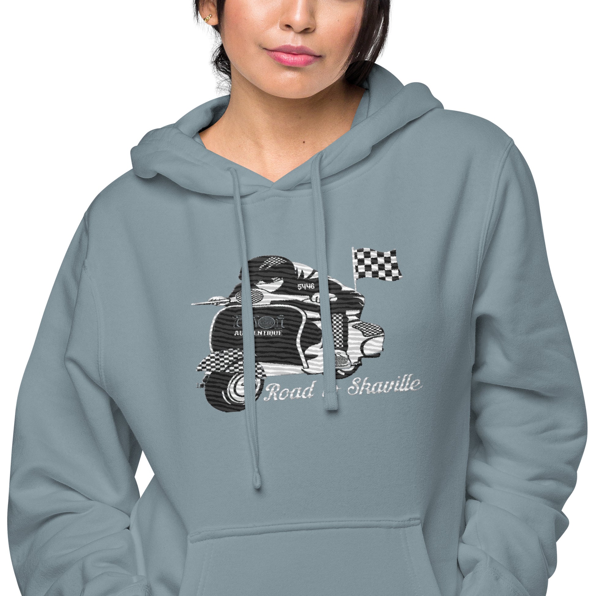 A woman wearing a slate blue pigment-dyed hoodie with front and sleeve embroidery featuring a vintage moped design and the text 'Road to Skaville'.